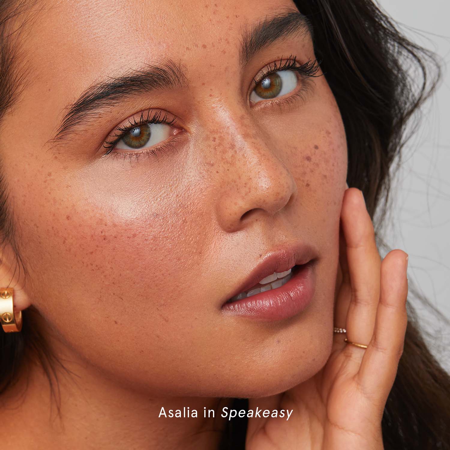 Close-up of a person with freckles touching their face and wearing a gold hoop earring.